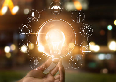 Strom 175896435 web Eine linke Hand hält eine leuchtende Glühbirne. Um die Glühbirne herum befinden sich Symbole in der Luft, die erneuerbare Energiequellen darstellen.